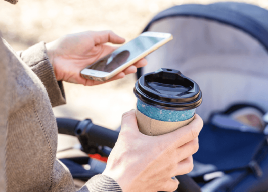 A woman pushes a pram while holding her mobile phone and a takeaway coffee cup.