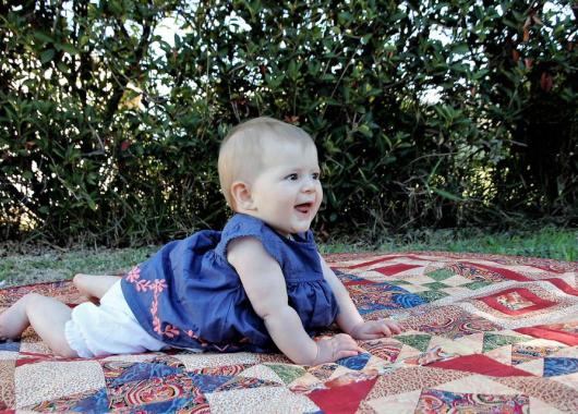 back ground shows trees. There is a colourful patchwork blanket on the grass. A baby girl is on the blanket. She is on her tummy and wears a blue top and white shorts. 