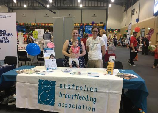 Two volunteers stand behind a table smiling. One has a baby in a carrier on her front