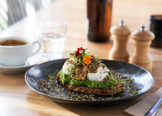 Toast with avocado, poached eggs and beautifully presented garnish is on a black plate. In the background is a cup of coffee, a glass of water and salt and pepper grinders. 