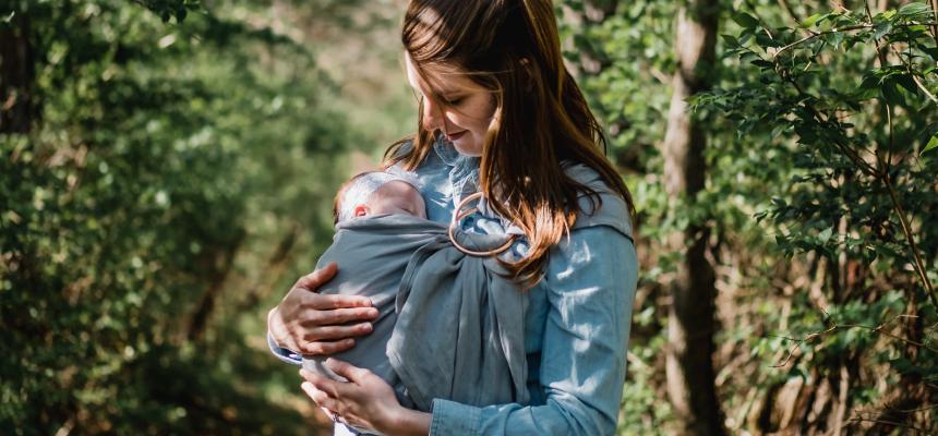 An image of a mum looking at the newborn baby she holds in a carrier, while standing outside in nature.