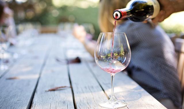 Red wine being poured from a bottle into a glass. The glass is on an outdoor table with people in the background at a party or celebration.