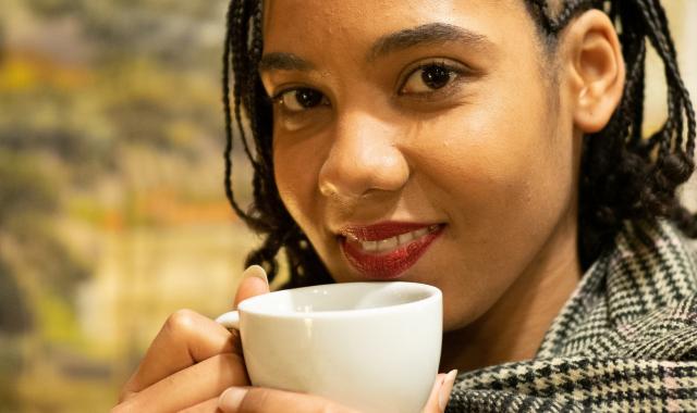 A woman is looking at the camera with a relaxed smile on her face. She has a mug cupped in her hands held up to her mouth. She's in the outdoors.