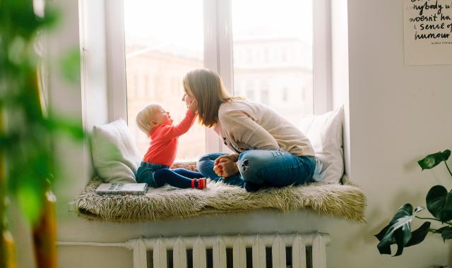 Mother and toddler playing together while sat in a window seat. Toddler is touching mum's face playfully..